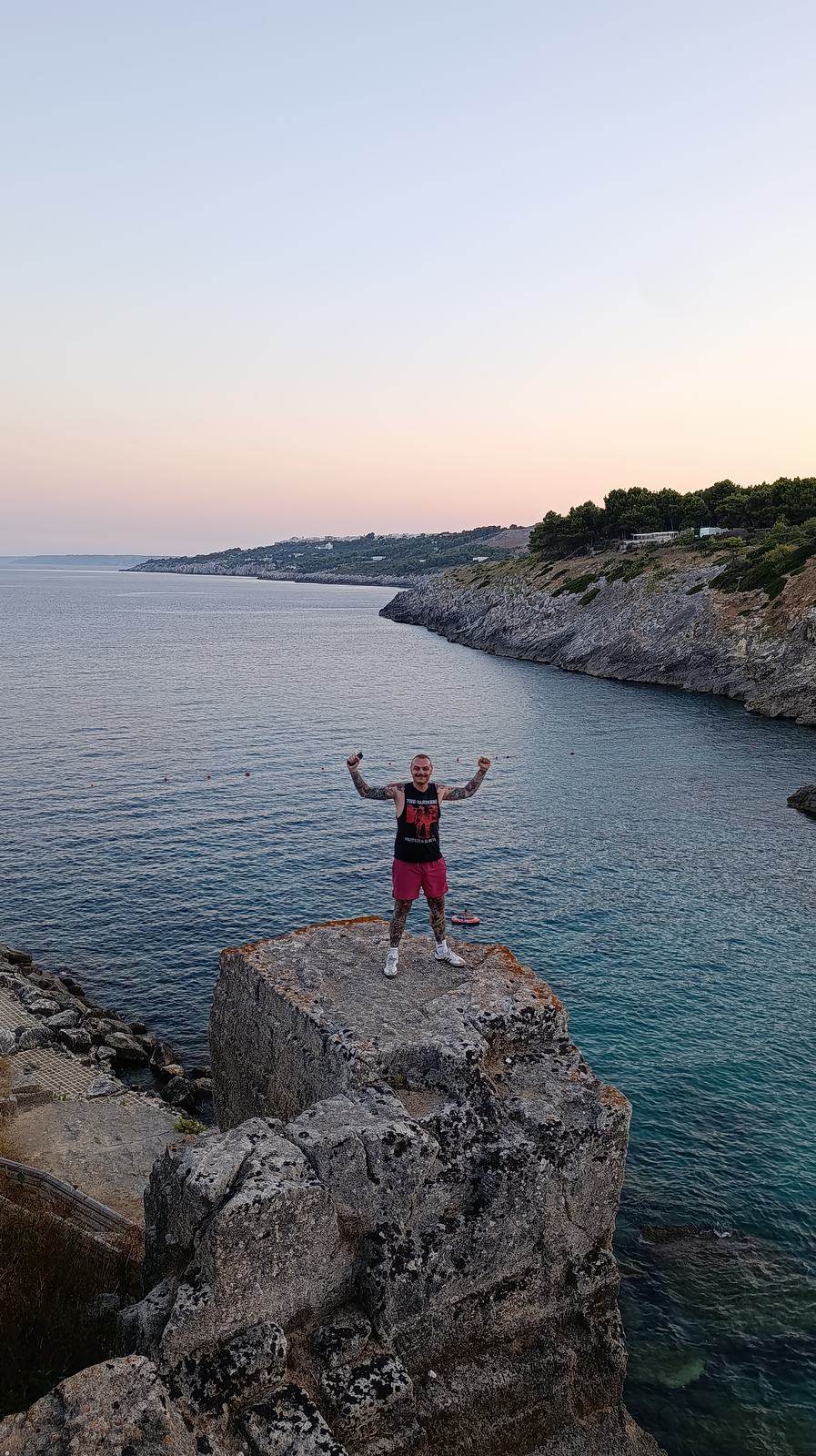 Moritz standing on a rock above the sea at sunset, arms raised, along the Salento coast