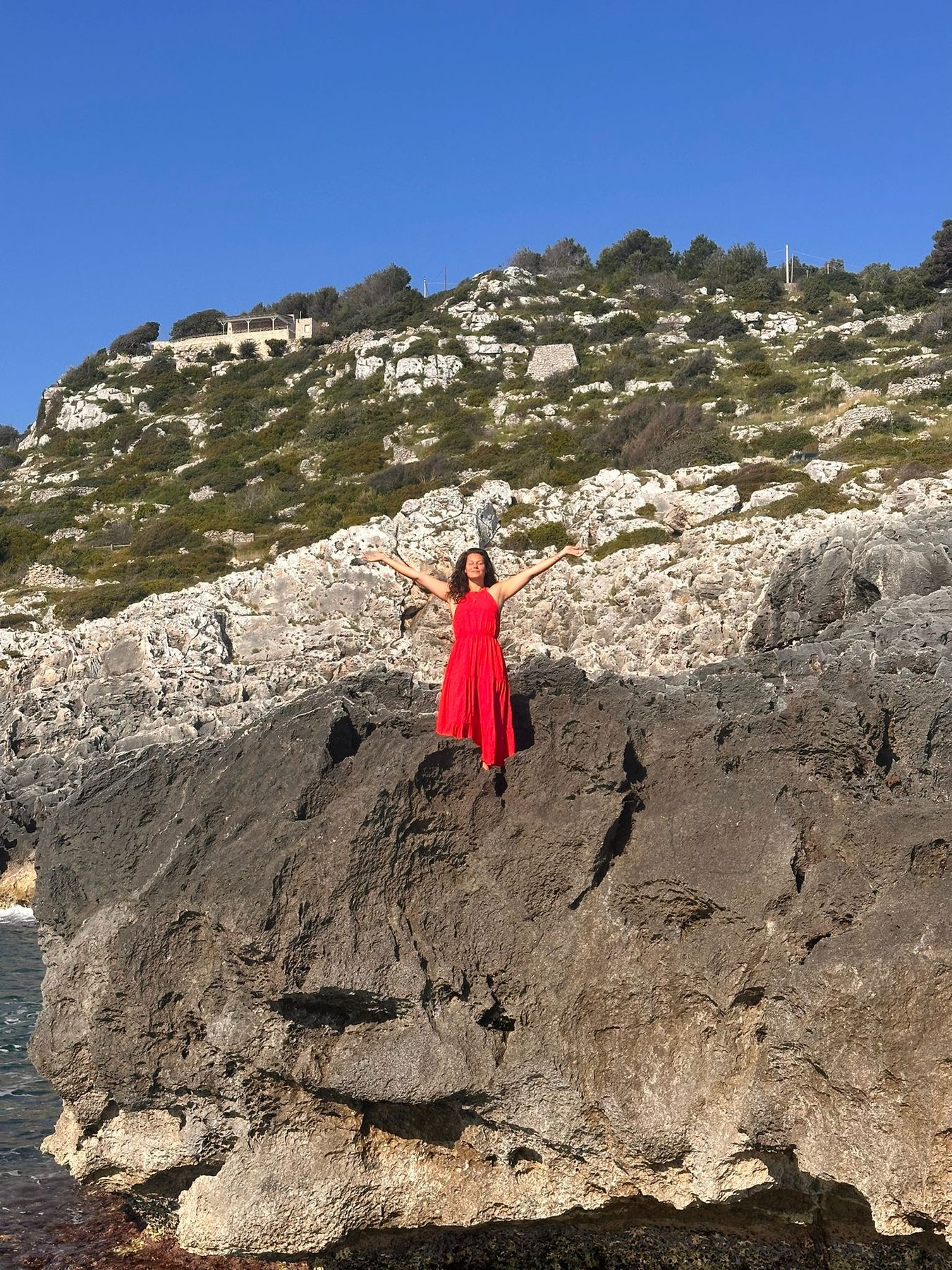 Nikita in red dress on Salento rocks with arms open wide