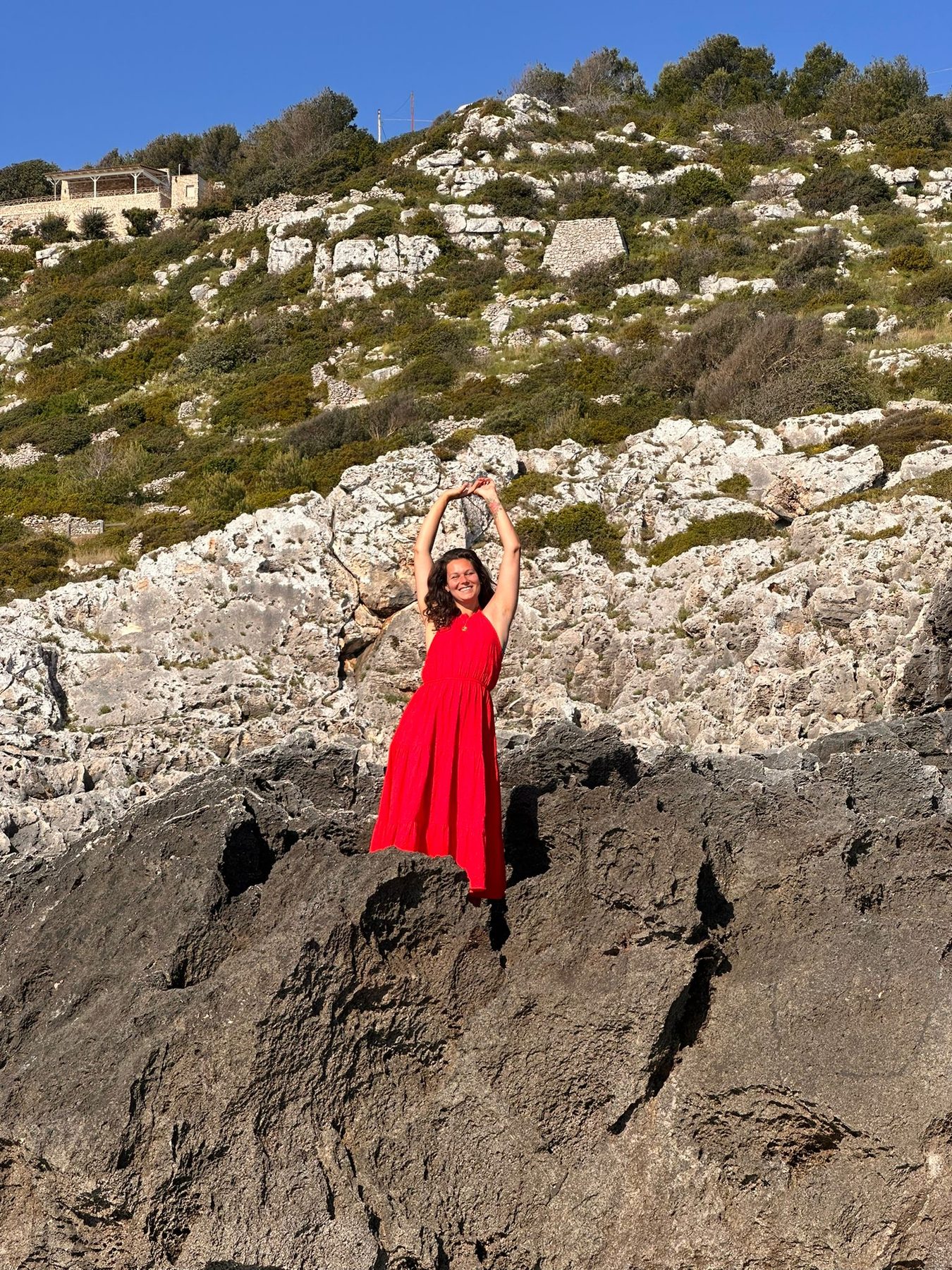 Nikita in red dress dancing on Salento rocks with arms raised