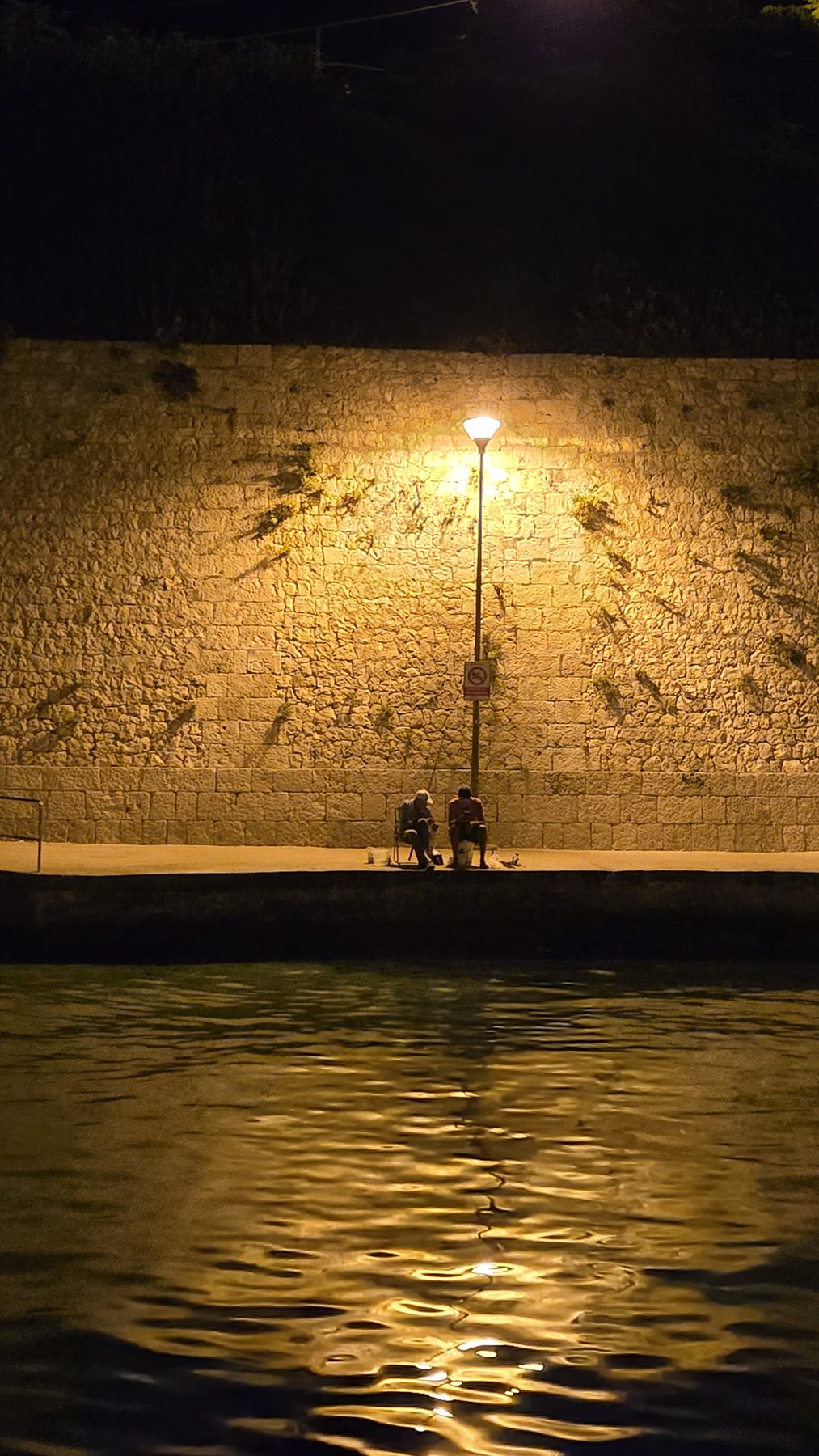 Two fishermen under a lamp on the harbour wall at Tricase Porto at night, with light on the water
